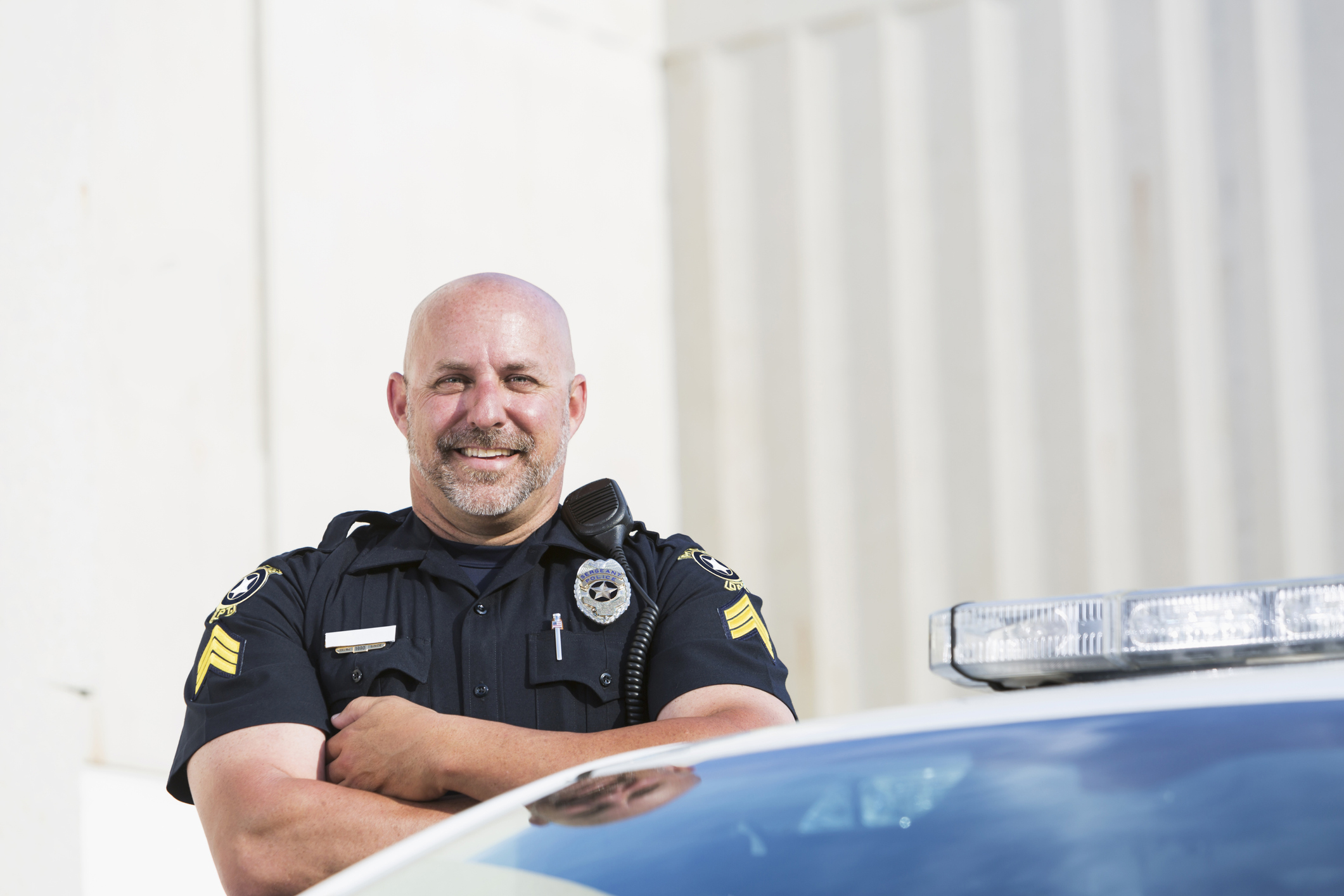 Policeman smiling, standing with arms crossed next to to his police car.   The man is in his mid 40s, heavyset and bald.  He is wearing a dark blue uniform.  Only the top of the cruiser, including the siren, is visible in the image.